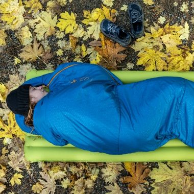 Person ligger i en sovepose p� et liggeunderlag i skoven omgivet af efter�rsblade, som illustrerer brug af soveposer og sovegrej i naturen.